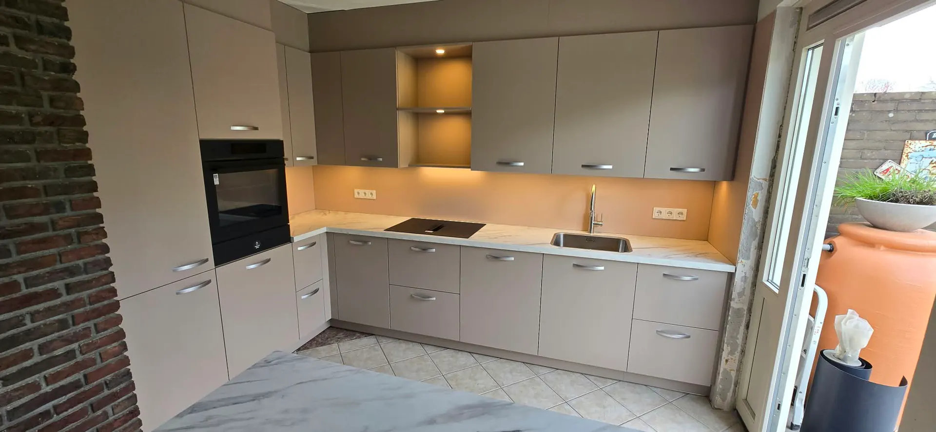 Lichte keuken met grijsbeige kasten, een warmgekleurde achterwand en verlichting onder de bovenkasten